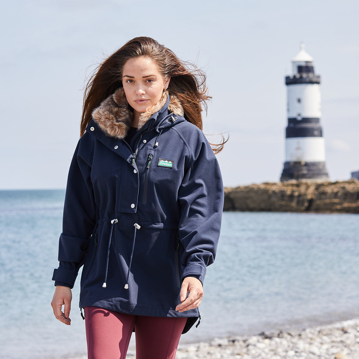 Woman wearing a navy blue coat with fur-lined hood on a beach with a lighthouse in the background