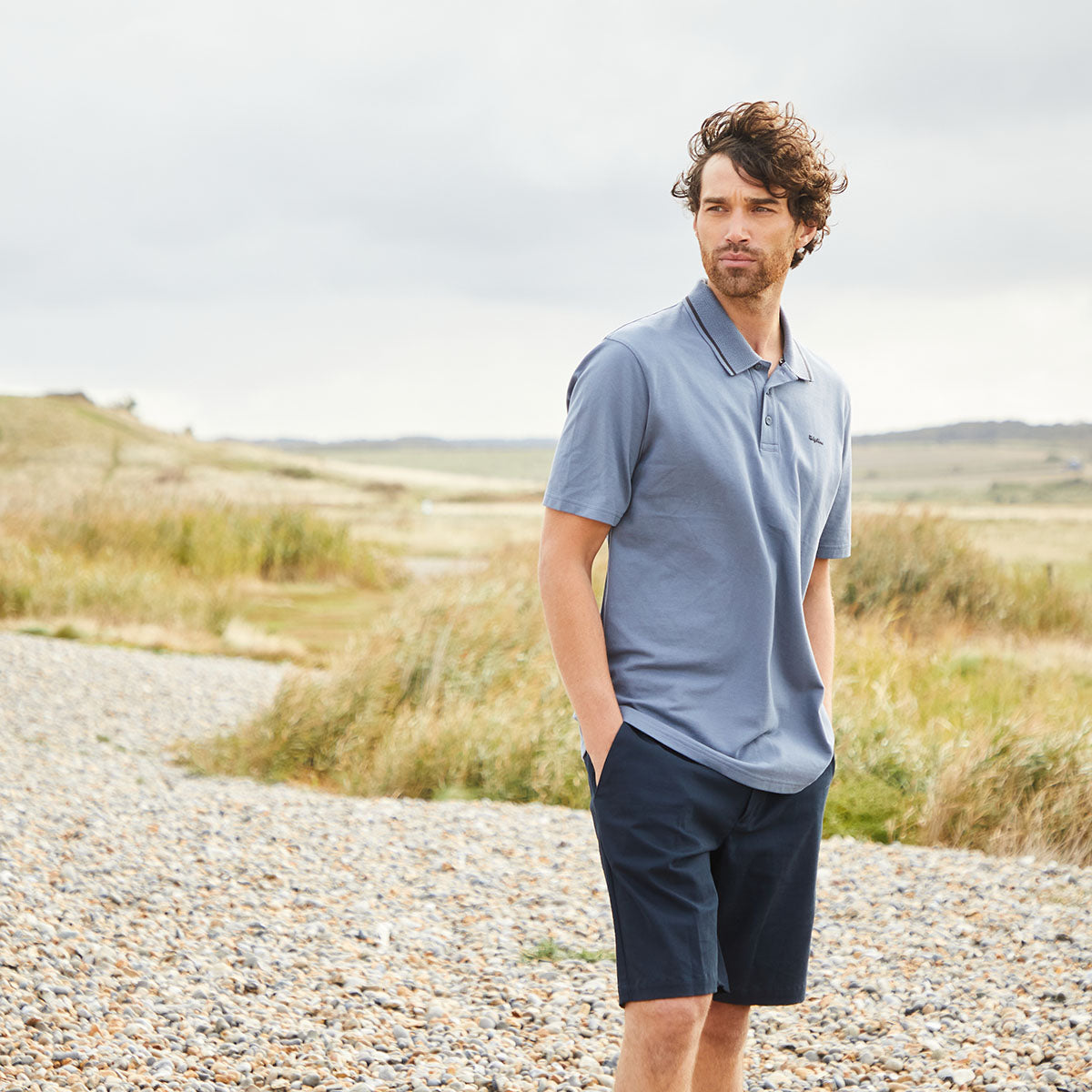 Man wearing Navy Chino shorts with Polo top standing on pebble beach