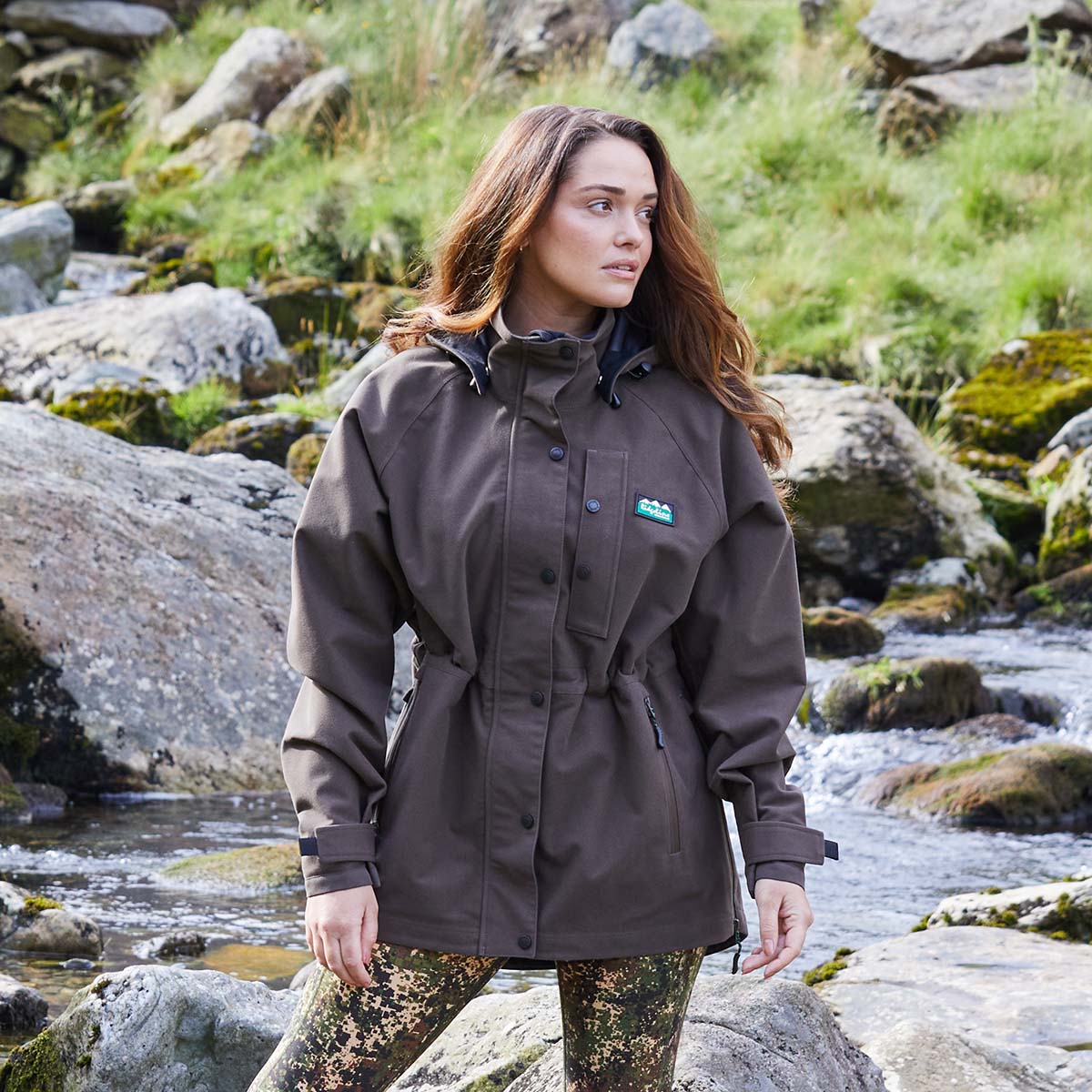 Woman wearing a brown waterproof jacket standing by a stream with rocks and greenery.