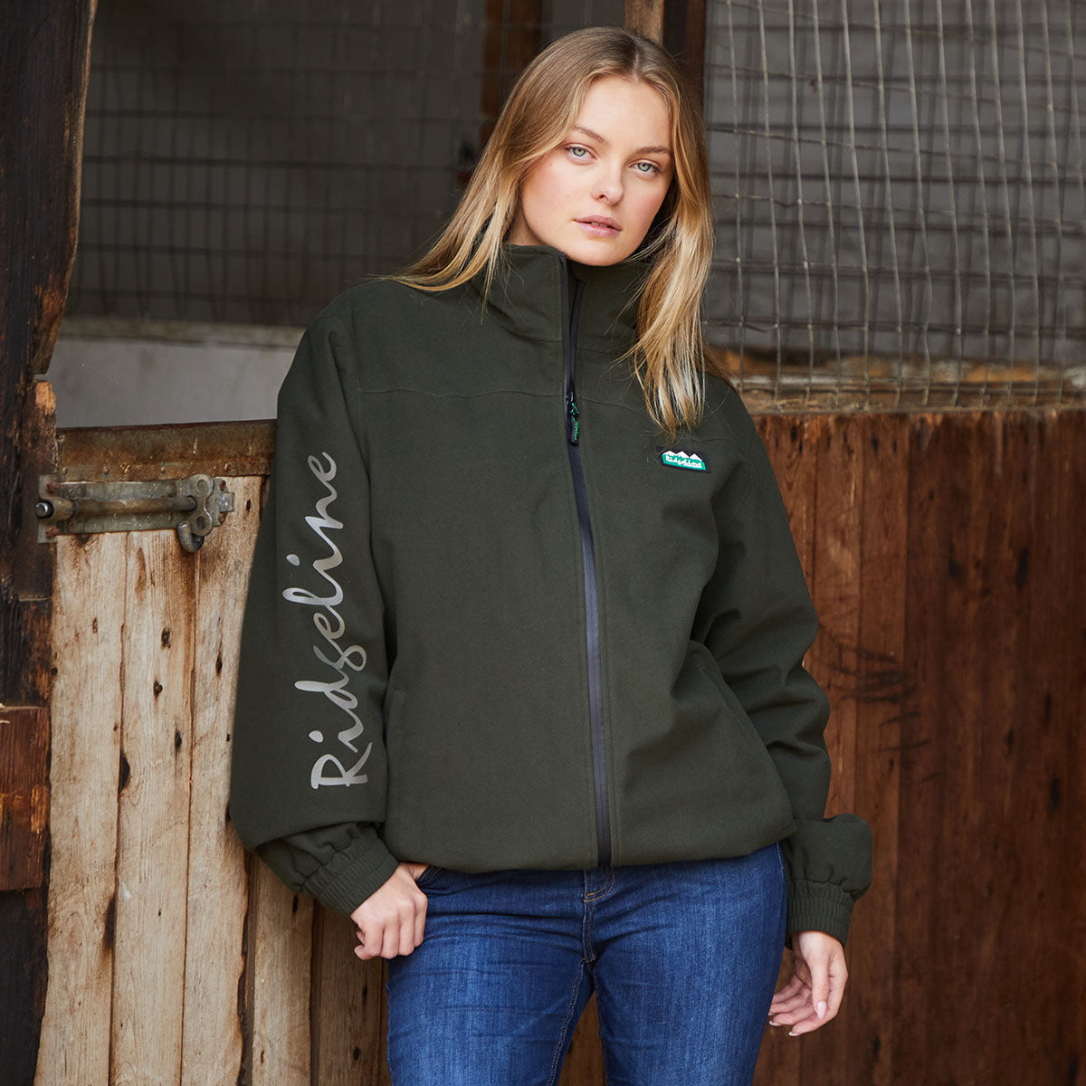 Woman wearing a green jacket with 'Ridgeline' branding in front of wooden stable.