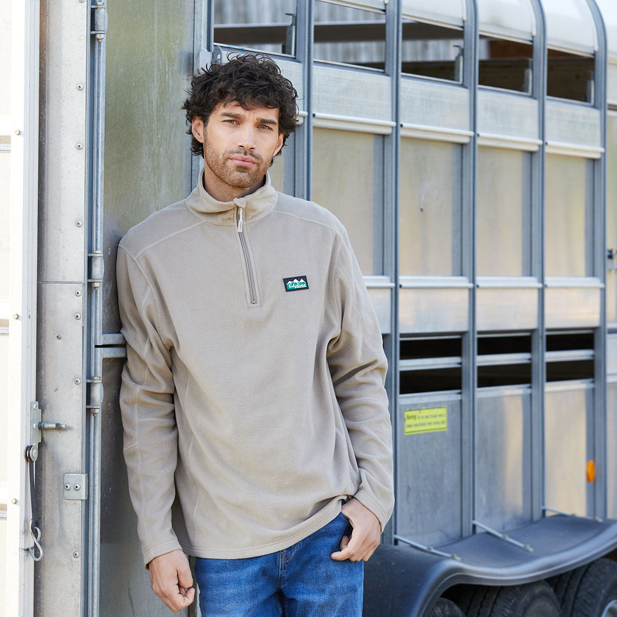 Man wearing a beige pullover with a Ridgeline Clothing logo standing next to a metal trailer.