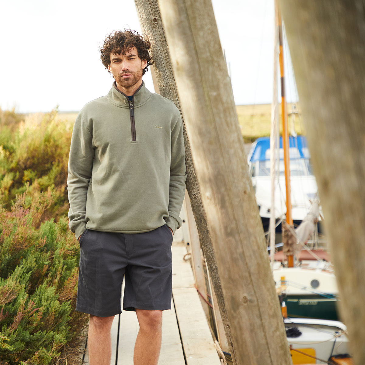 Man wearing black chino shorts with quarter zip jumper standing on boardwalk next to boats