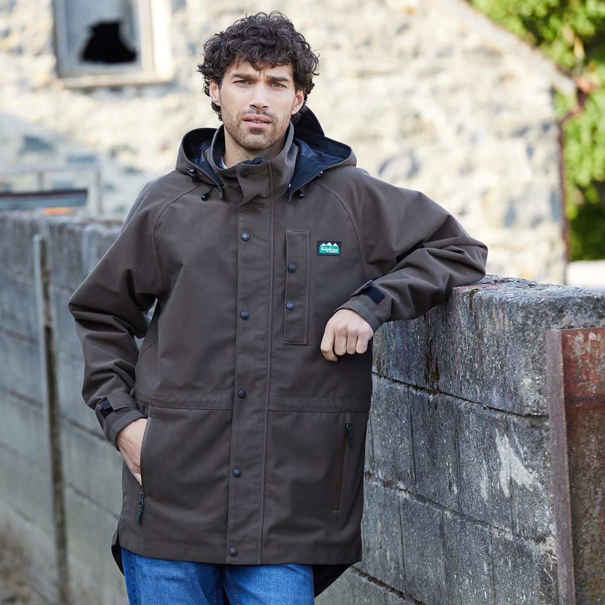 Man wearing a brown waterproof jacket with a Ridgeline Clothing brand logo, leaning against a stone wall.