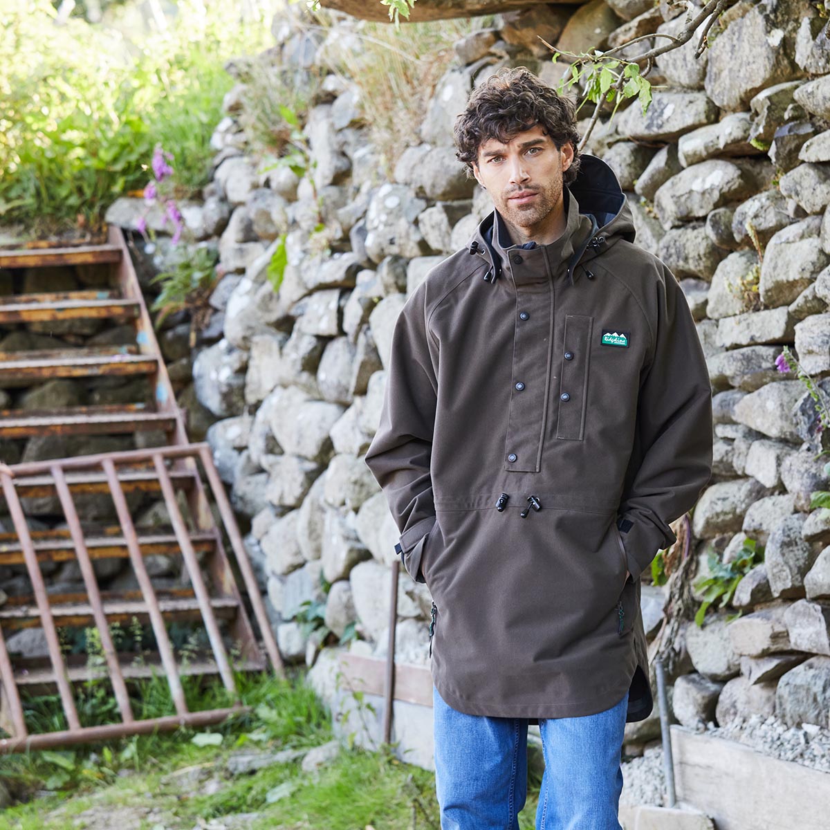 Man wearing a brown waterproof jacket with a logo standing in front of a stone wall.