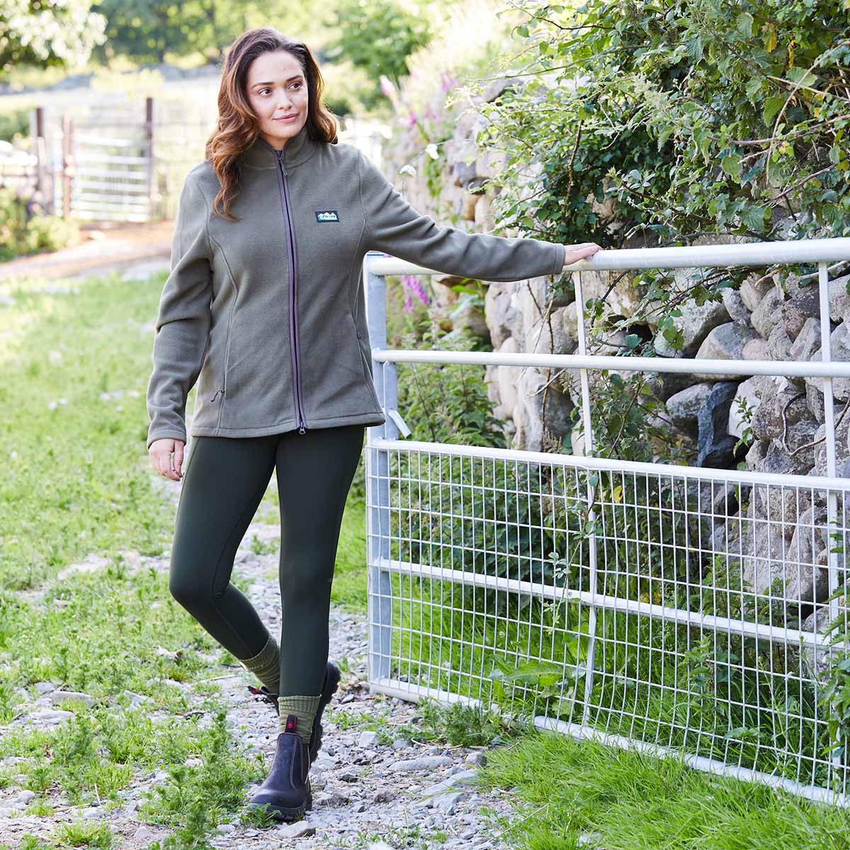 Woman in a green jacket wearing green leggings standing next to a metal gate in a garden setting