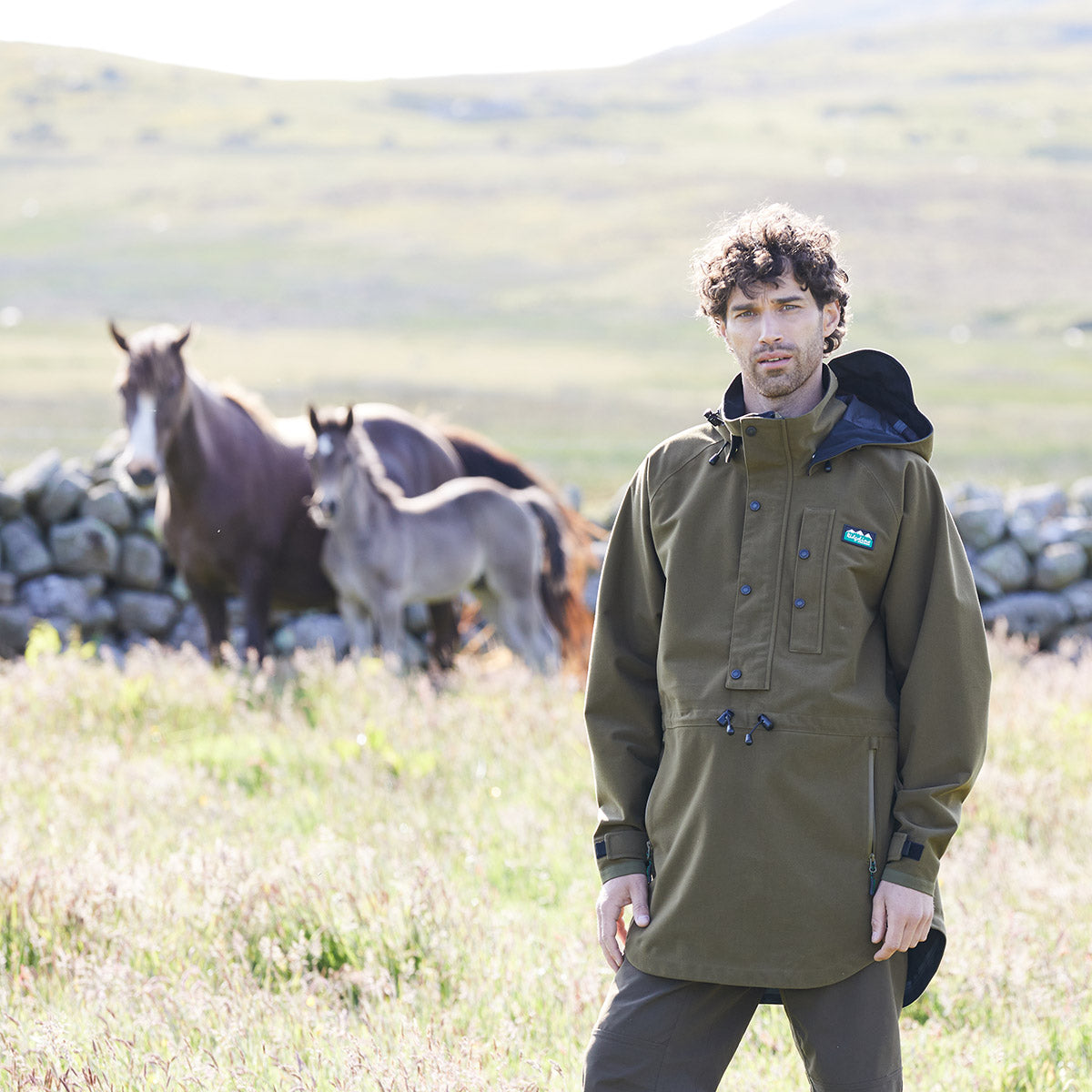 Man wearing a brown waterproof jacket standing in a field with horses in the background