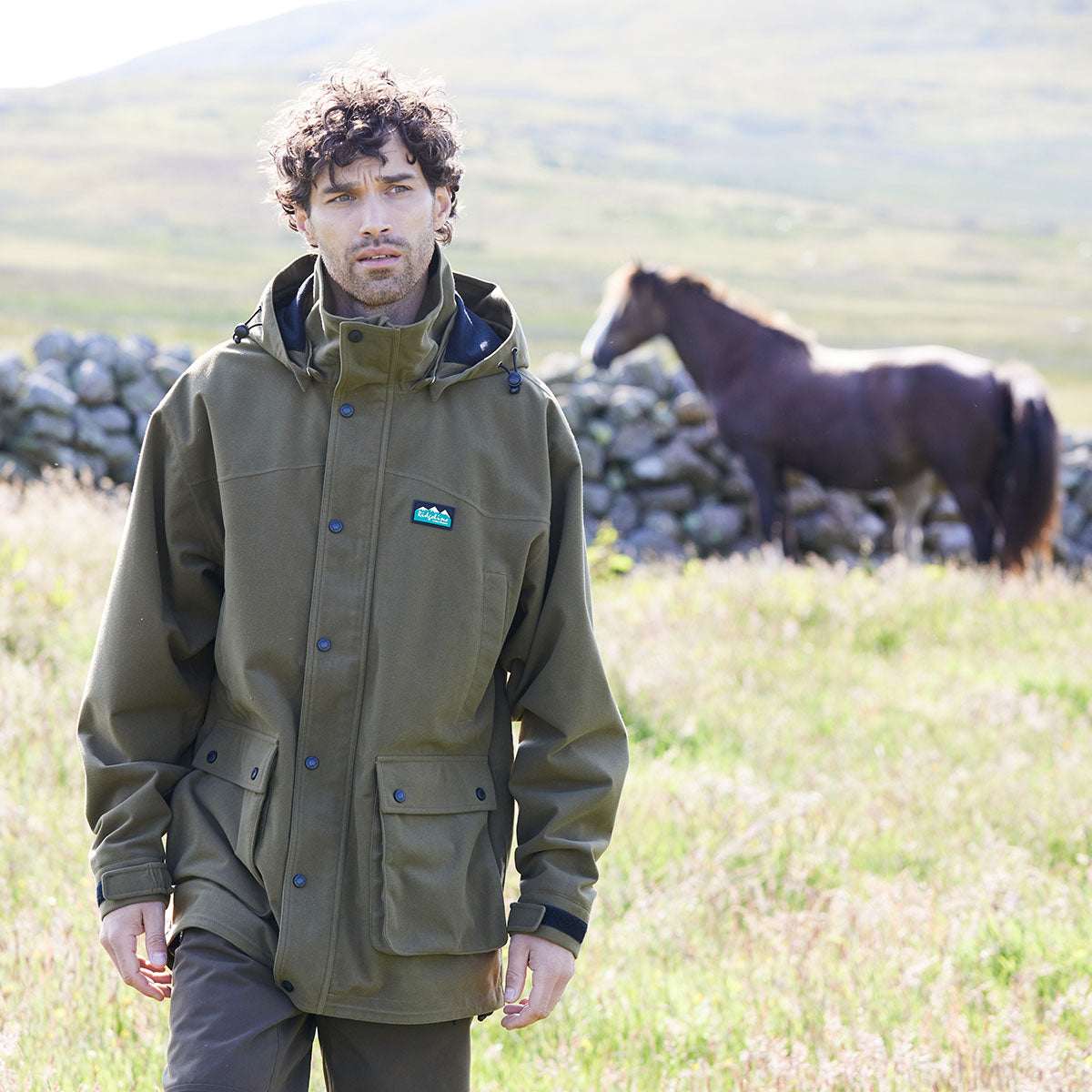 Man wearing a brown waterproof jacket standing in a field with a horse in the background