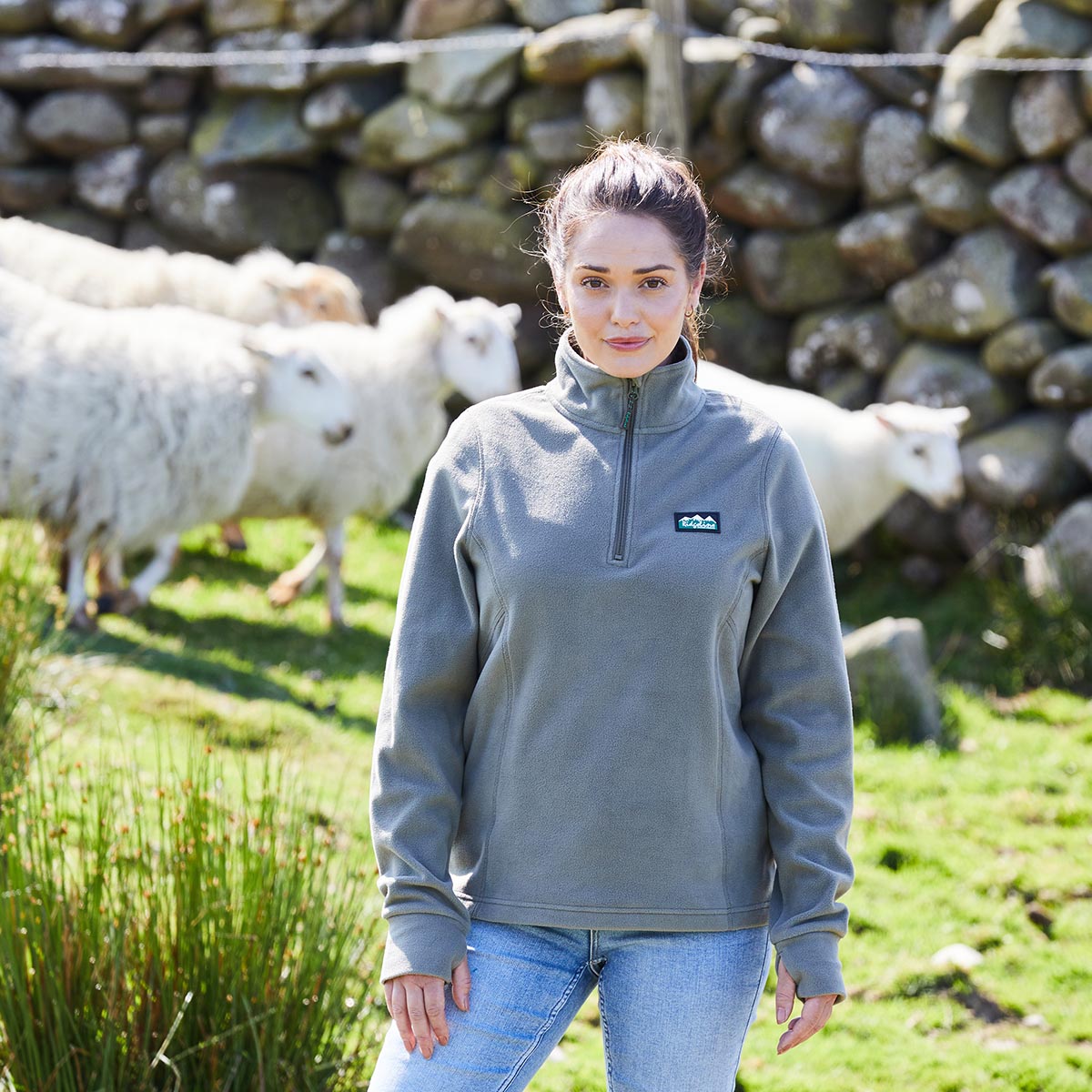 Woman wearing a gray fleece with a Ridgeline Clothing Logo, standing in a field with sheep and a stone wall in the background.