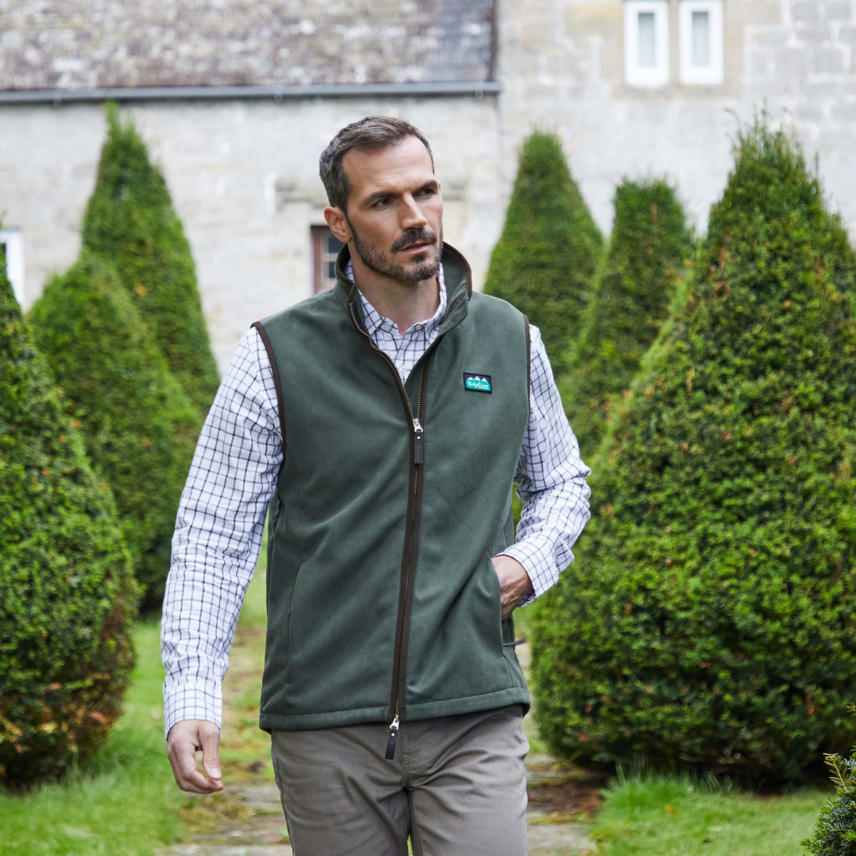 Man wearing a green vest over a checkered shirt in front of a building with green bushes.