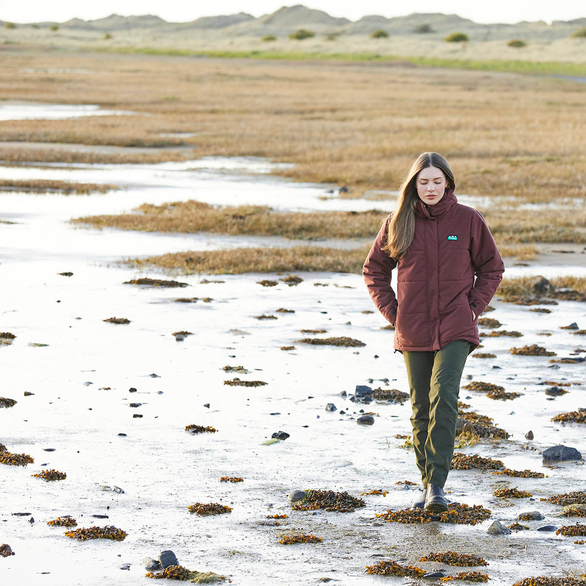Person wearing a Ridgeline insulated jacket in burgundy with a high collar and side pockets, walking across a coastal shoreline with seaweed-covered sand and shallow water in the background.