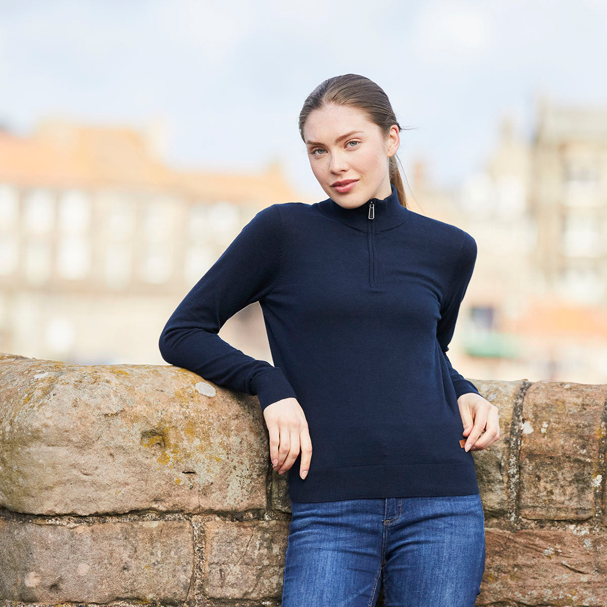 Person wearing a navy blue quarter-zip knit sweater with a high collar and long sleeves, paired with blue jeans, leaning against a stone wall outdoors. The sweater features a fine-knit construction, ribbed hem and cuffs, and a streamlined fit. The image is photographed in a natural setting with a softly blurred background of buildings, highlighting the sweater's style, fit, and everyday wear versatility.