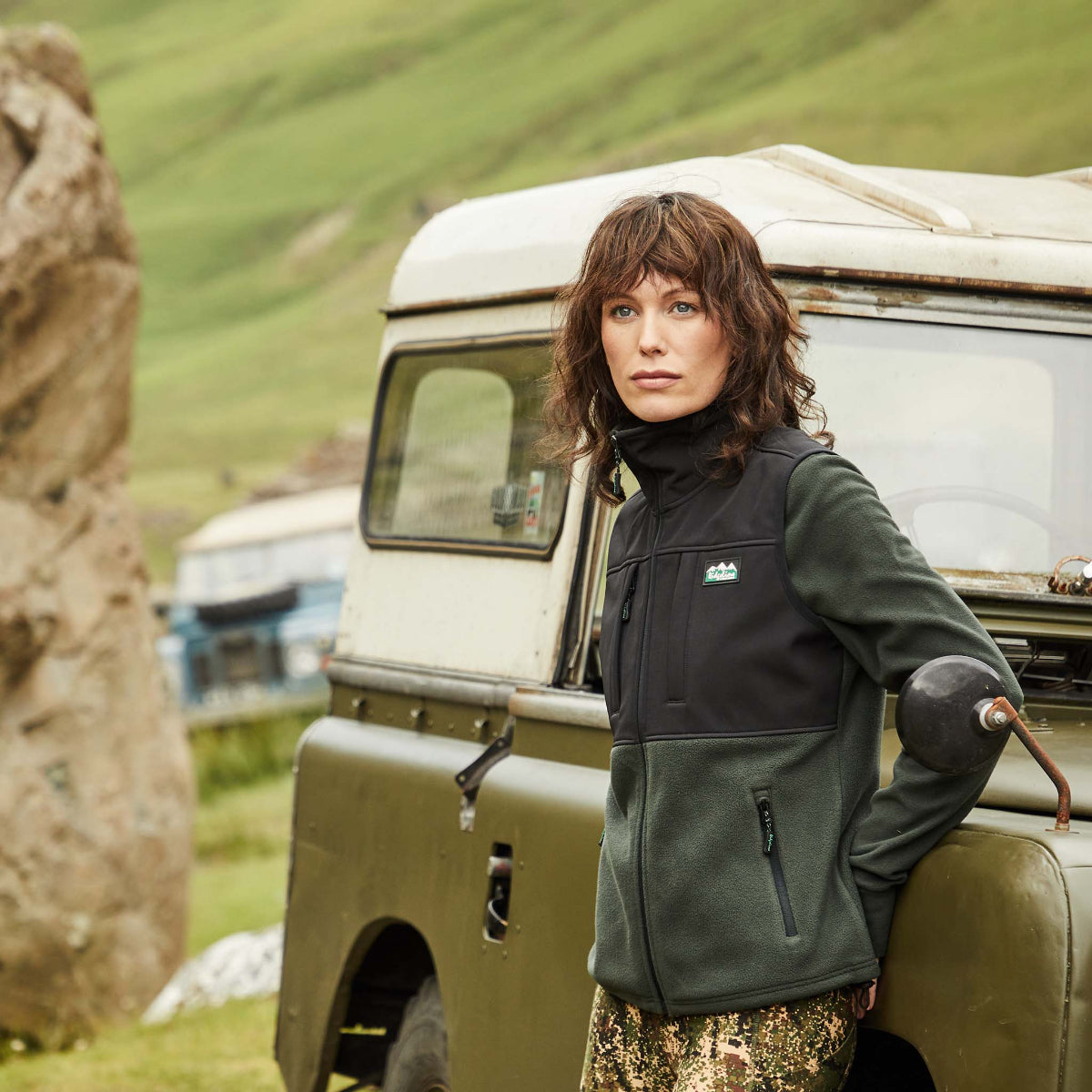 a women wearing a black and green hybrid vest with a full-length front zip, chest pockets with a land rover in the background.