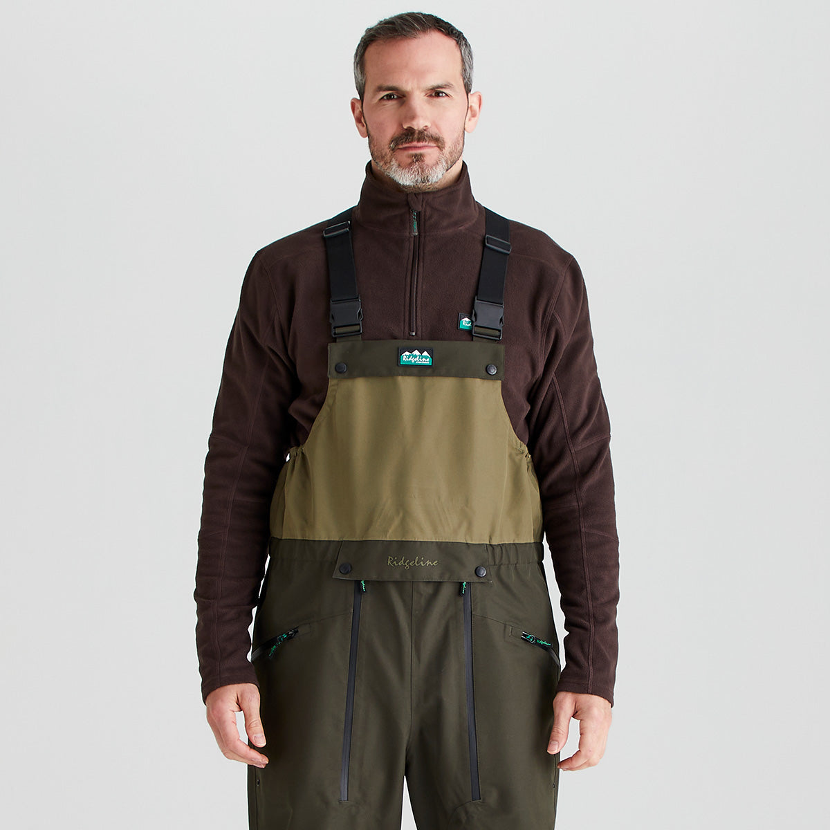 Man wearing a brown jacket and green waders/ bib & brace on a light gray background