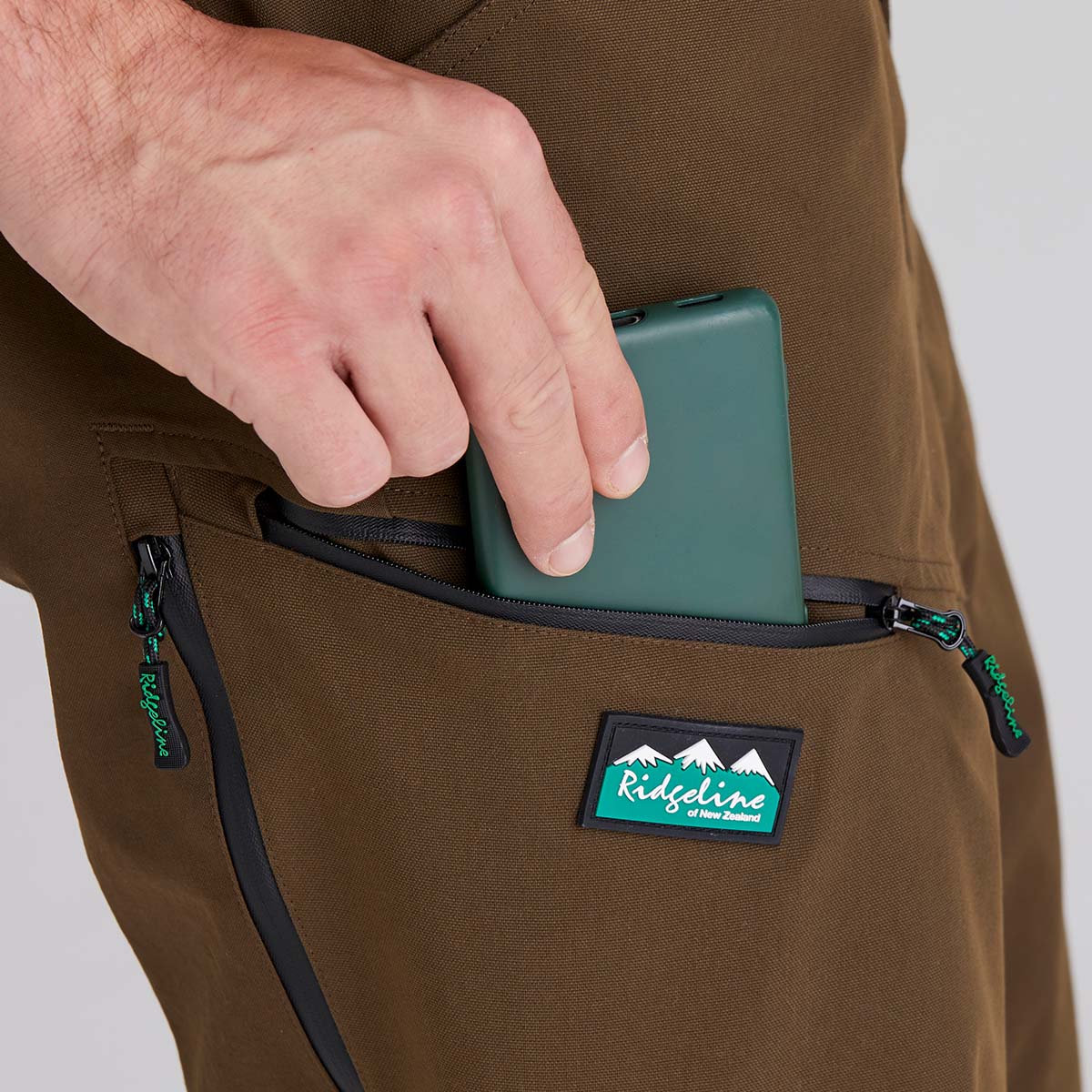 Person inserting a green device into a pocket of brown pants with a Ridgeline logo.