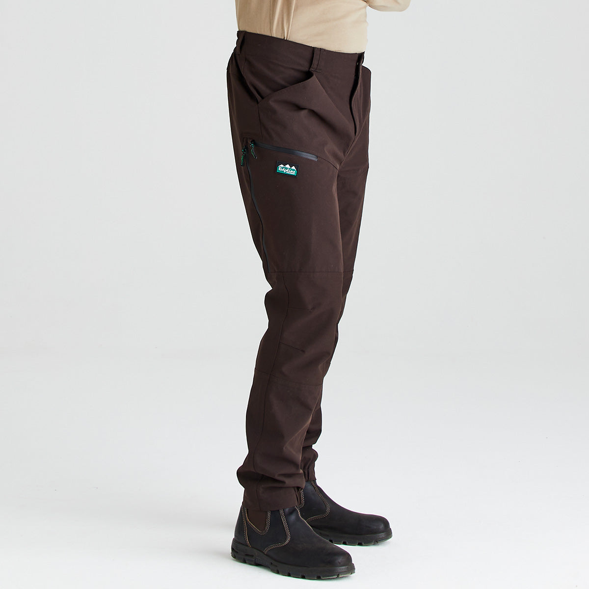 Side view of dark brown work trousers worn with black work boots, shown against a light grey studio background. The trousers feature a straight-leg fit, articulated knee panels, side hand pockets, and a zippered utility pocket on the thigh with a small logo detail. A beige long-sleeve top is partially visible above the waistband.