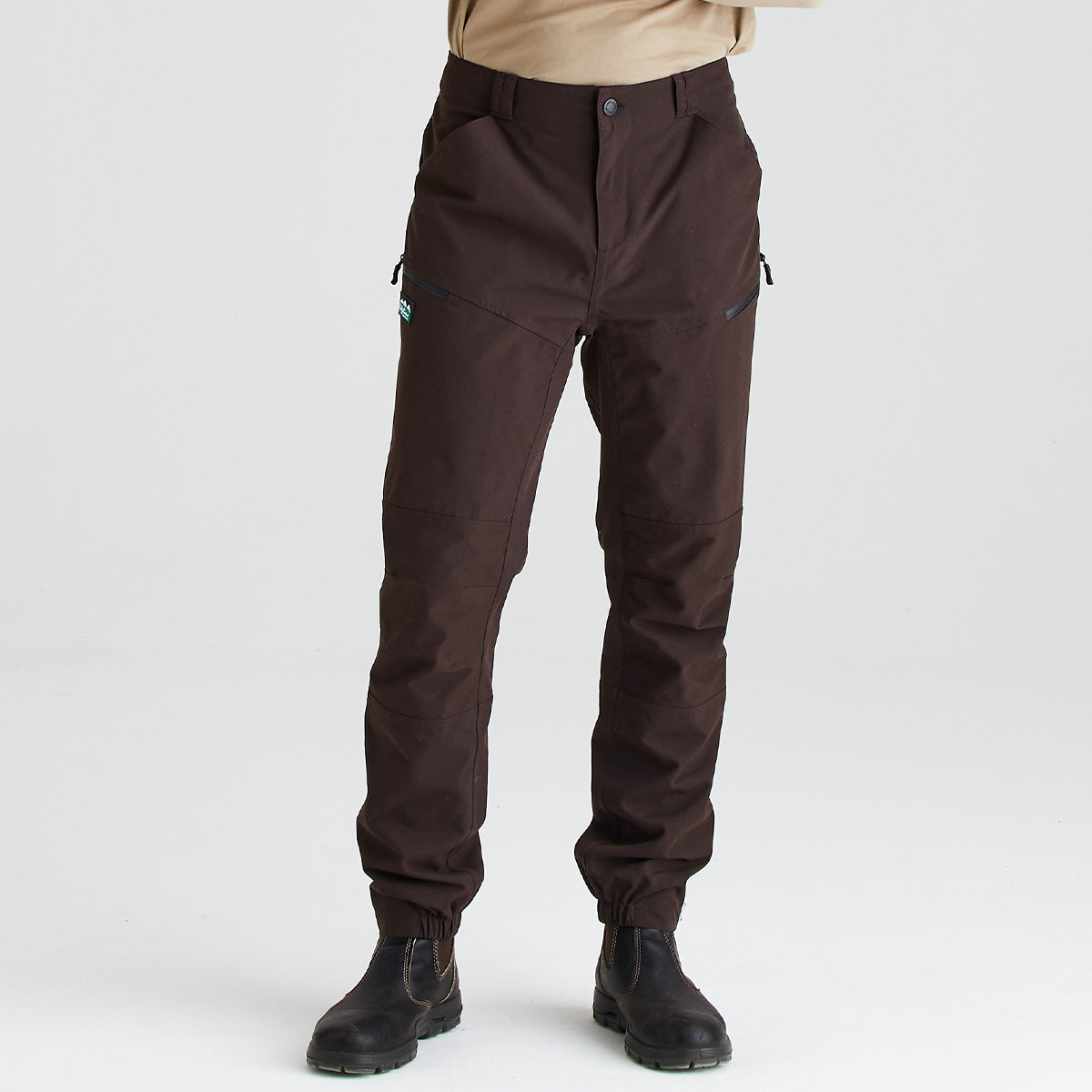 Dark brown outdoor work trousers shown from the waist down on a model against a light grey background. The trousers feature a straight-leg fit, reinforced knee panels, belt loops, side hand pockets, and zippered utility pockets on the thighs. Black work boots are worn with the trousers.