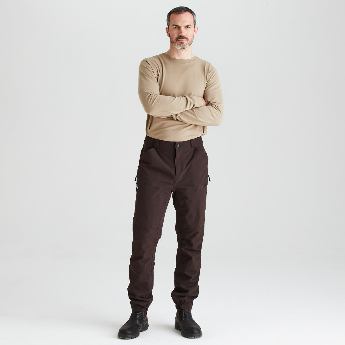 Full-length studio image of a person wearing dark brown work trousers with reinforced knee panels, zippered thigh pockets, and a straight-leg fit. The trousers are paired with a beige long-sleeve top and black work boots. The person is standing with arms crossed against a light grey background.