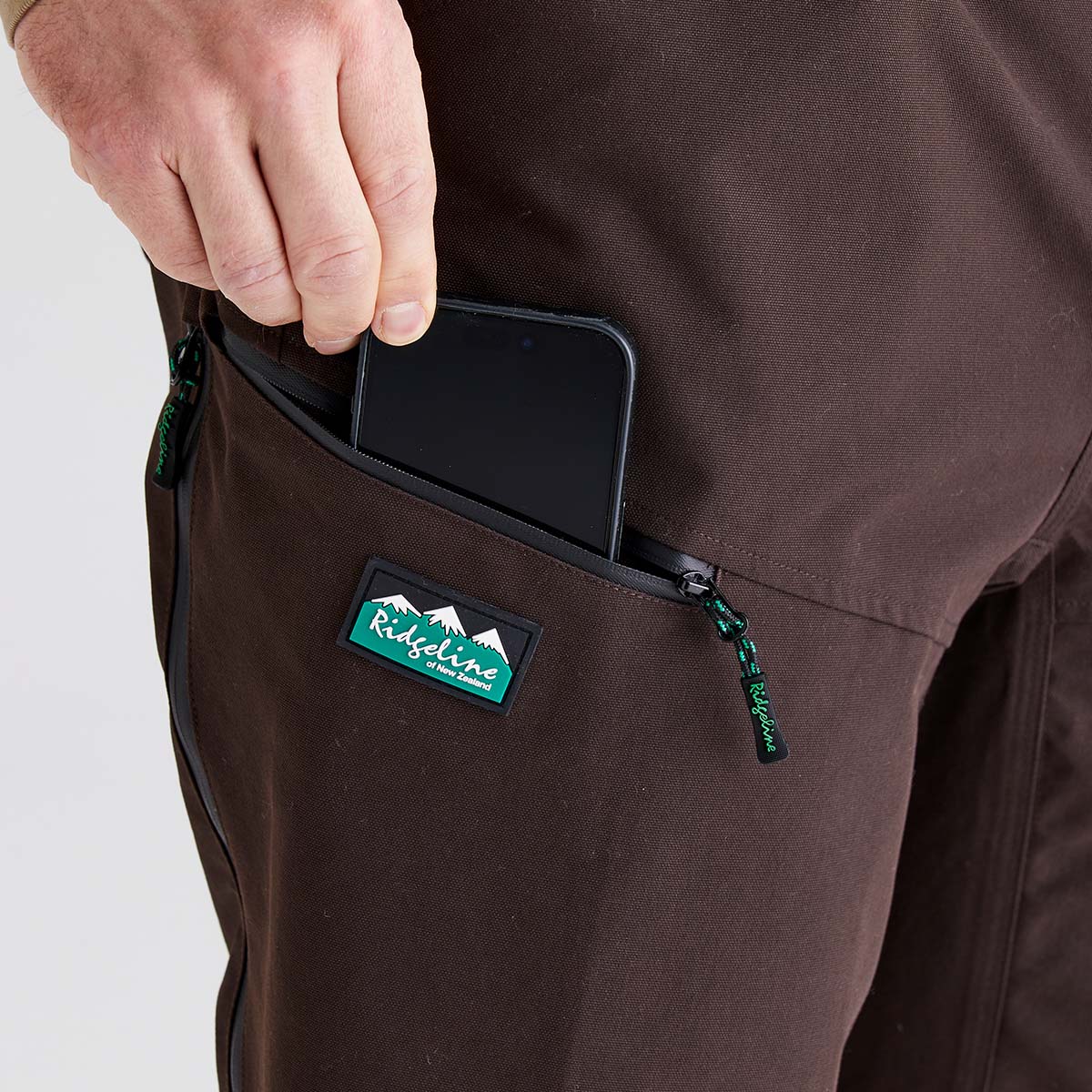Close-up of brown work trousers featuring a zippered thigh pocket. A hand is placing a black smartphone into the pocket, demonstrating the pocket’s storage capacity. The trousers have black zippers with green pull tabs and a visible Ridgeline logo patch on the pocket. The background is plain white.