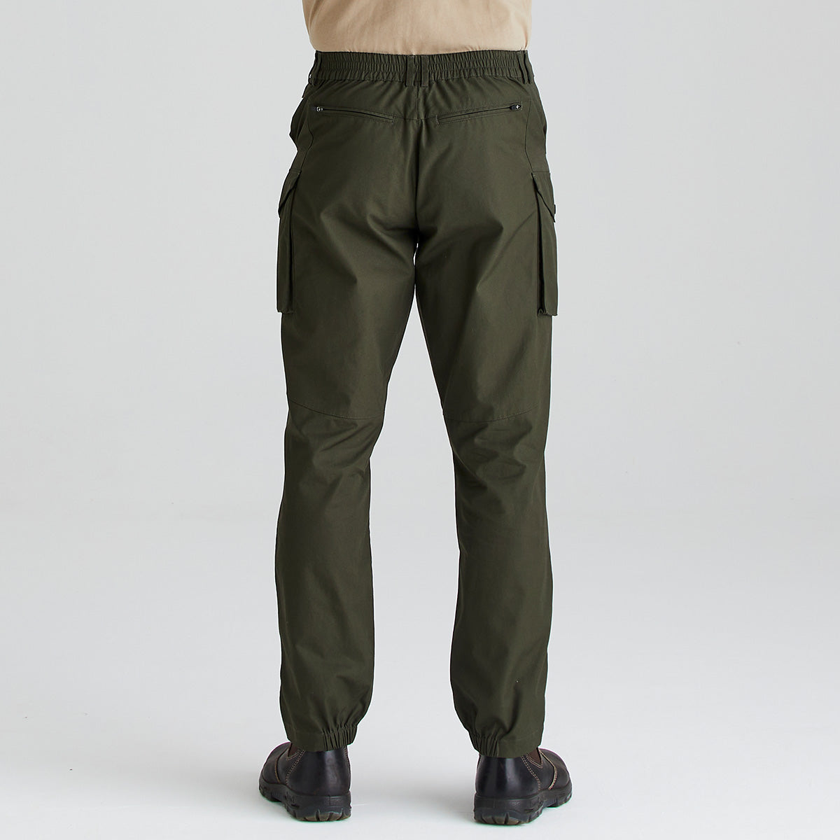 Rear view of olive-green cargo work trousers worn with dark leather work boots and a beige long-sleeve top. The trousers feature an elasticated waistband at the back, straight legs with elasticated ankle cuffs, zipped rear pockets, and large cargo pockets on both sides. Shown against a plain light-grey studio background, the image highlights the back fit and functional storage design of the trousers.