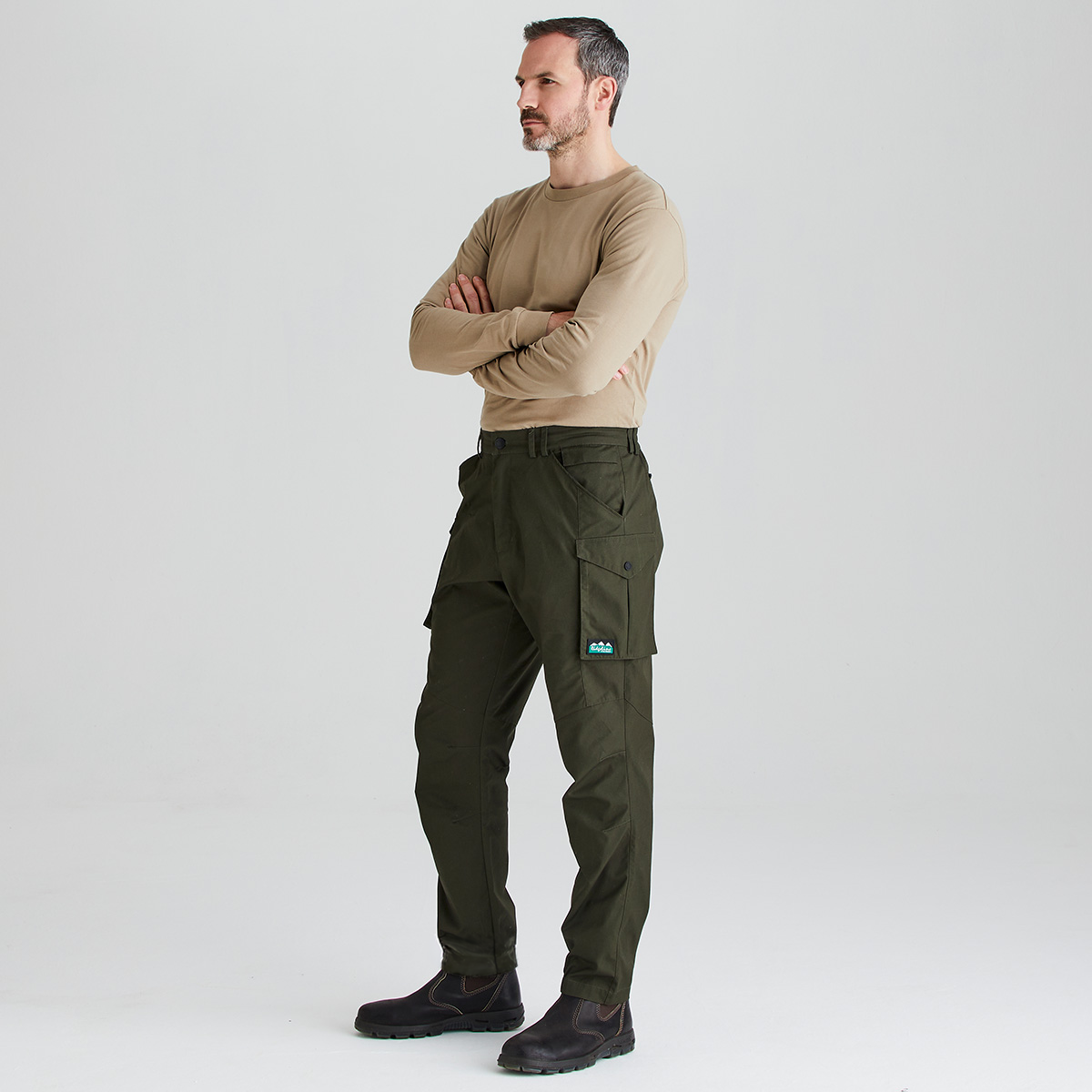 A man modeling dark olive green cargo pants with a tan long-sleeve shirt, standing against a light gray studio background.