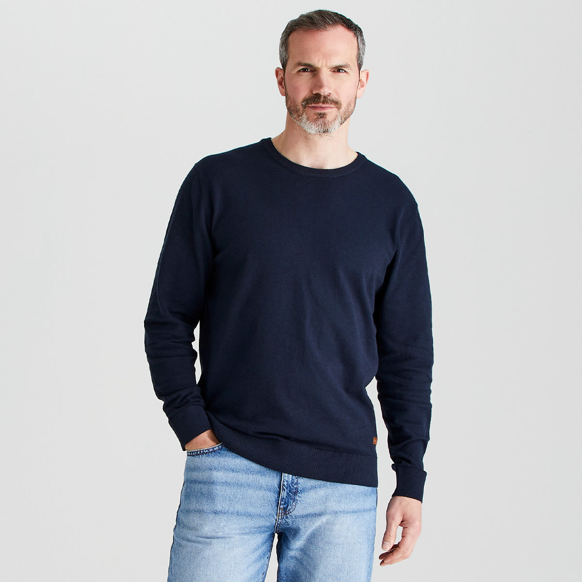 Man wearing a navy sweater and light blue jeans on a plain background