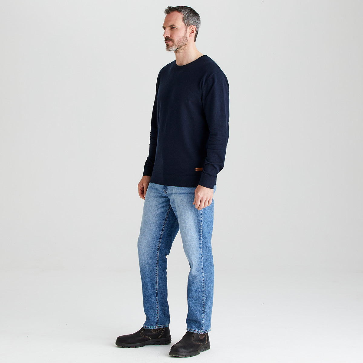 Man wearing a navy long-sleeve jumper and blue jeans on a white background