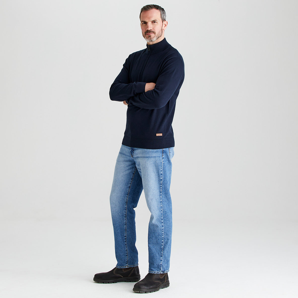 Man wearing a navy sweater and blue jeans standing against a light gray background