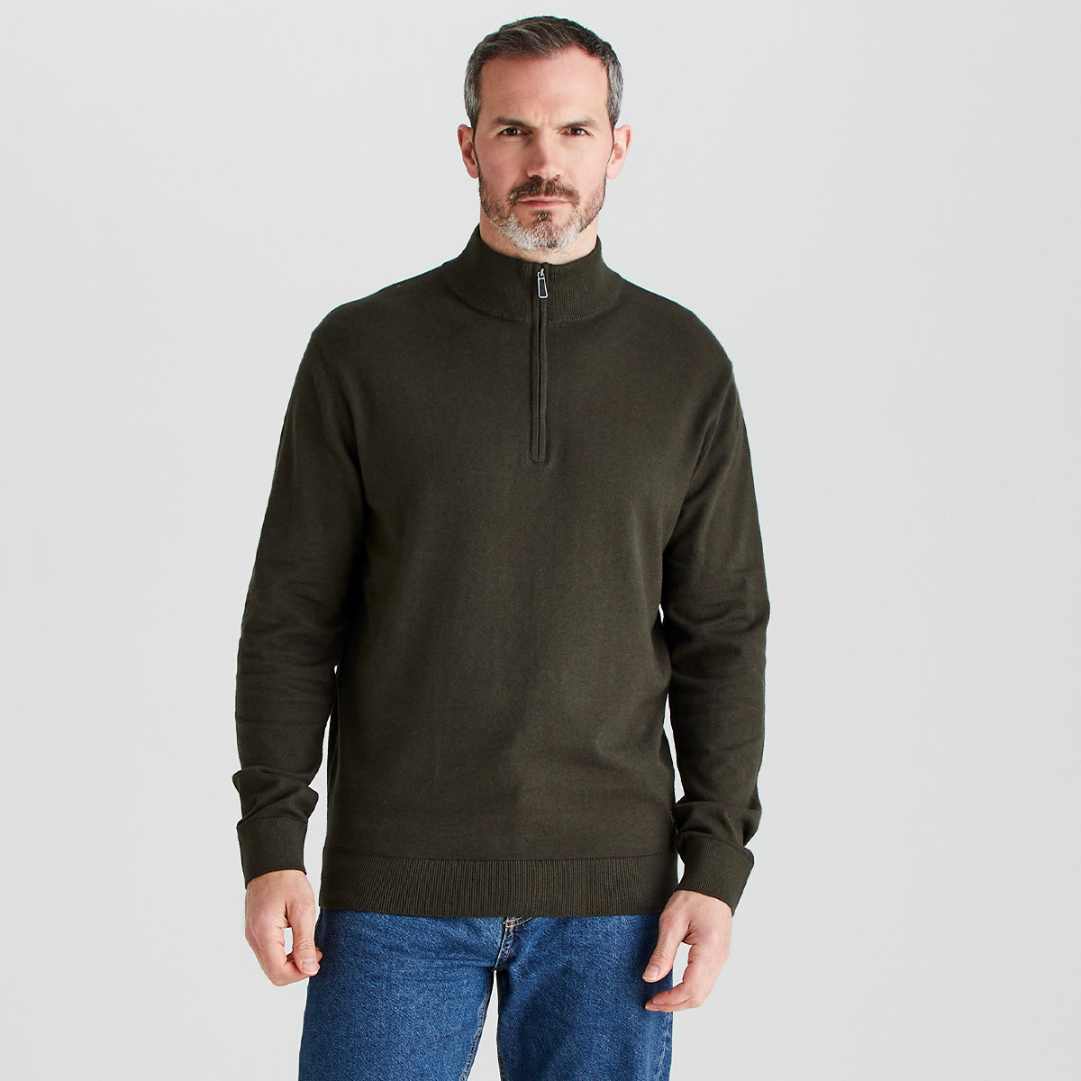 Man wearing a dark green half-zip sweater and blue jeans on a light gray background