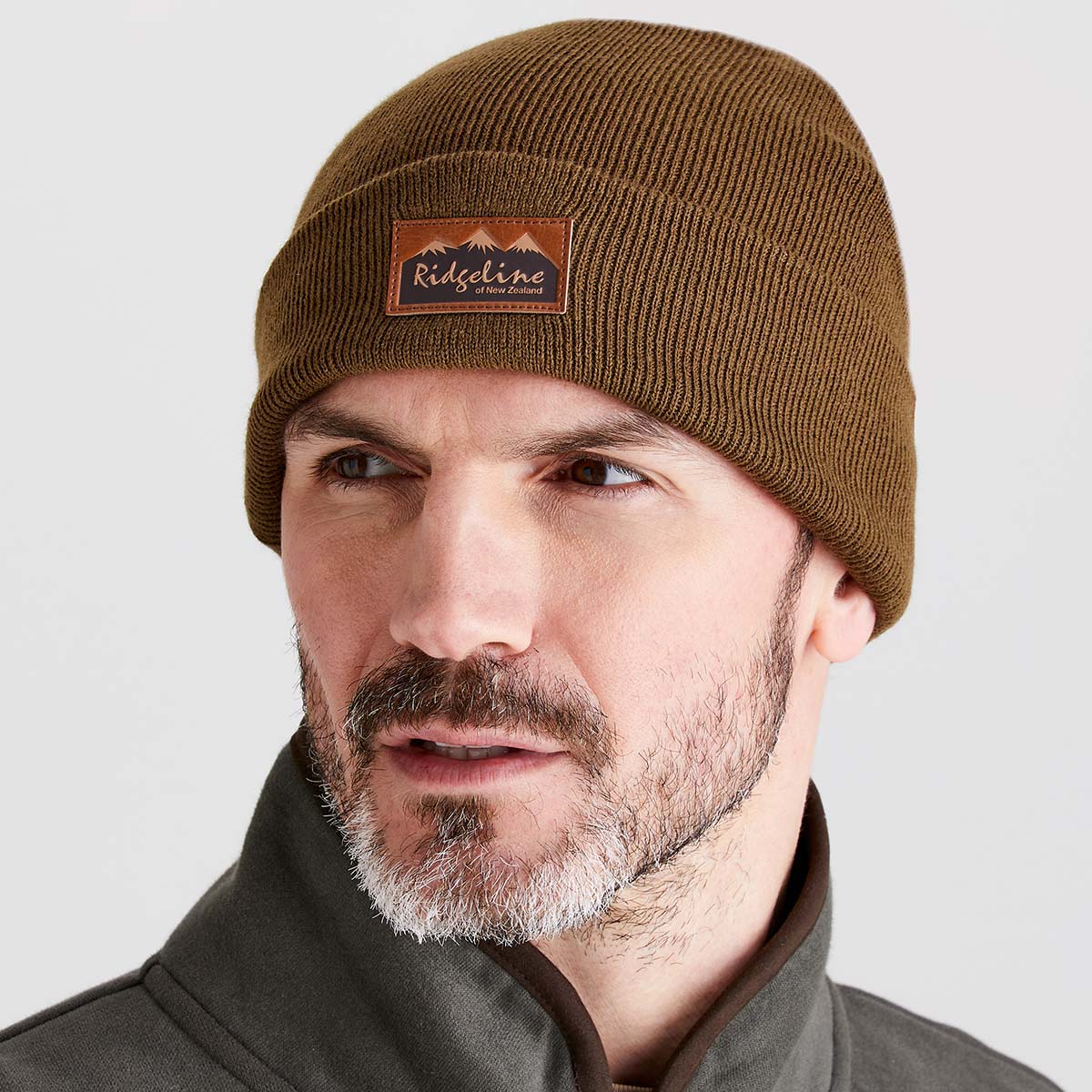 Close-up lifestyle image of a person wearing a greenish-brown ribbed knit beanie with a folded cuff. A brown faux-leather patch on the front displays the Ridgeline of New Zealand logo with a mountain graphic. The beanie is paired with a dark outdoor jacket and shown against a light grey background, highlighting the knit texture, snug fit, and classic cold-weather styling.