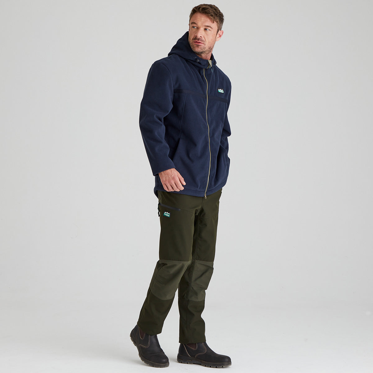 Man wearing a navy windproof fleece jacket and green pants on a light grey background