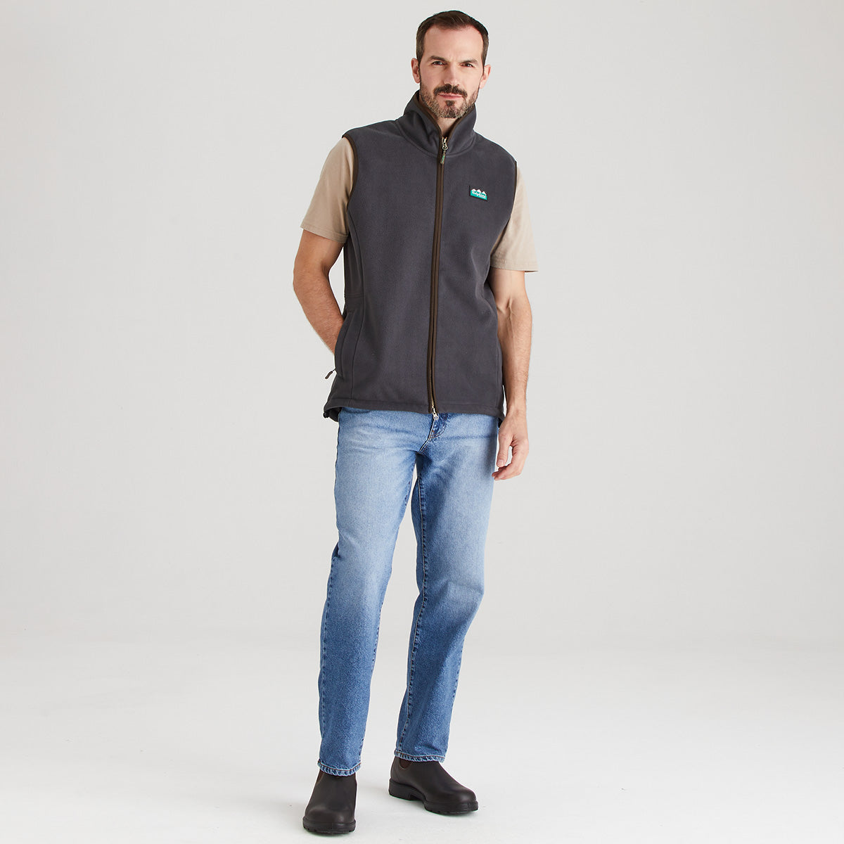 Man wearing a gray vest over a beige shirt with blue jeans on a white background