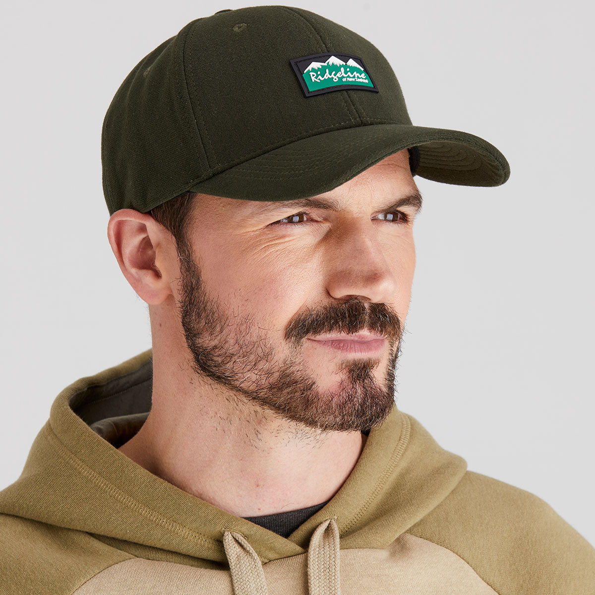 Man wearing a green cap with a logo and a beige hoodie on a light gray background