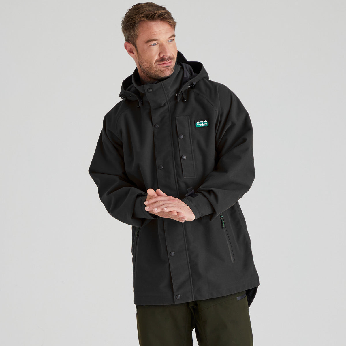 A man wearing a black hooded outdoor jacket with a small green and white logo on the left chest, paired with dark olive pants, photographed against a light gray studio background.