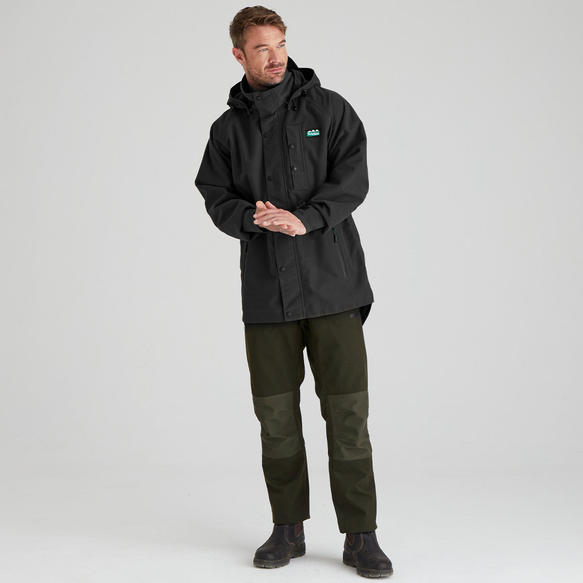A man wearing a black hooded waterproof jacket with a small green logo on the left chest, paired with olive green pants and black boots, standing against a light gray studio background.