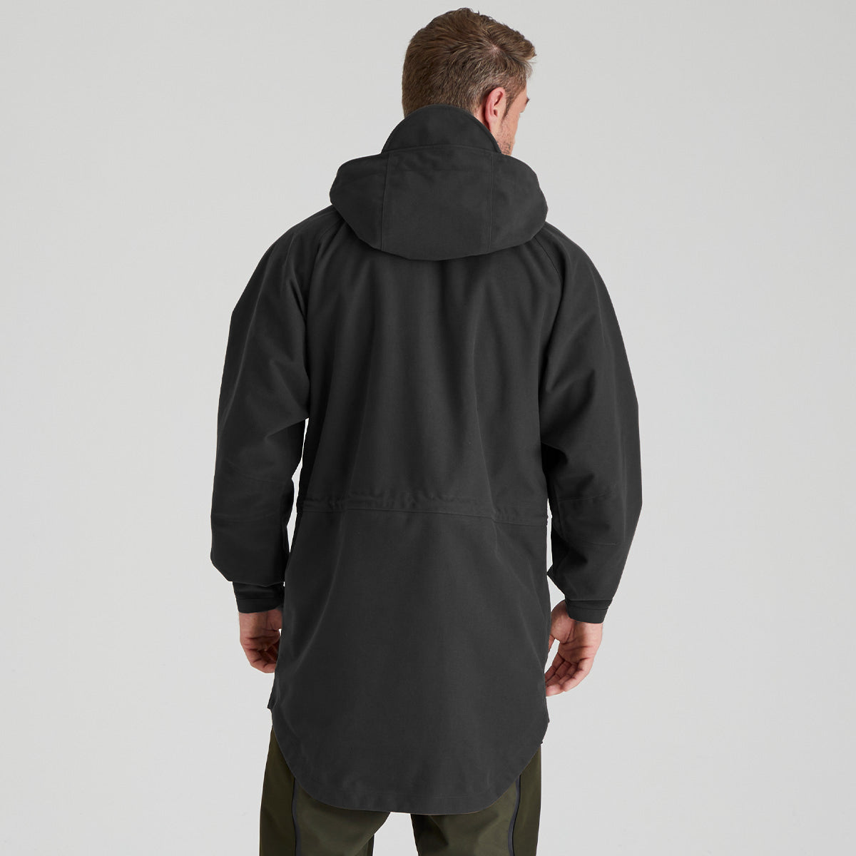 A man viewed from behind wearing a black hooded jacket and olive green pants against a light gray studio background.