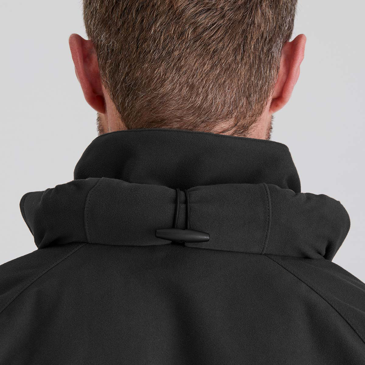 Close-up back view of a man wearing a black high-collar jacket with a toggle closure, set against a plain light gray background.