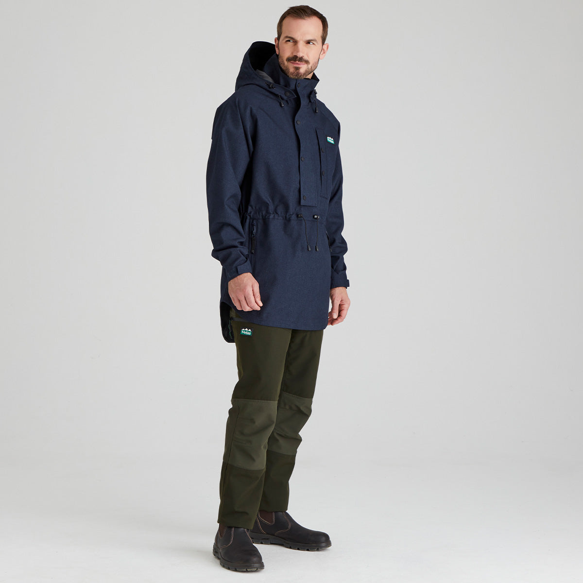 Man wearing a navy blue waterproof jacket and green pants on a light grey background