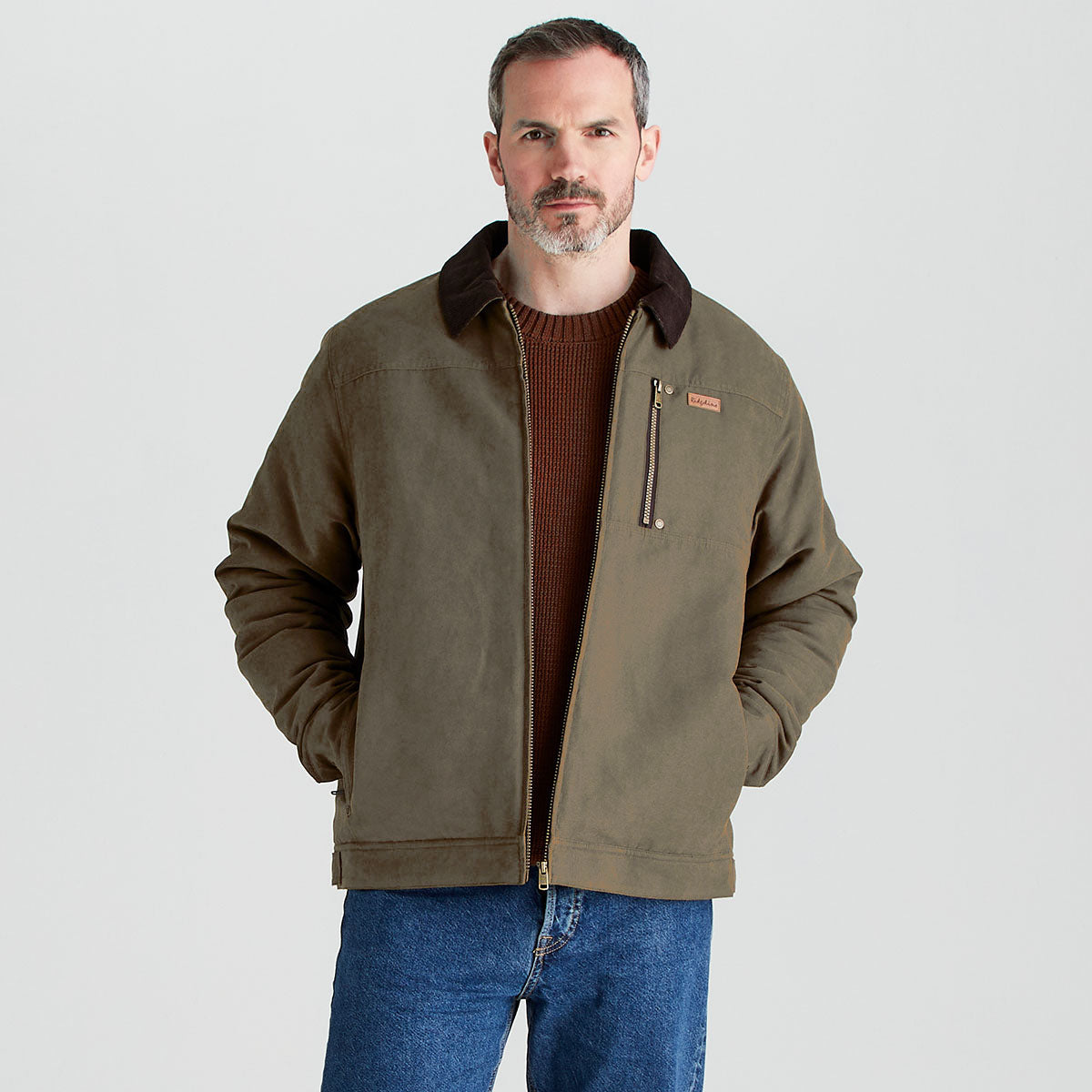 Front-facing studio image of a person wearing an olive-green zip-up jacket with a dark brown contrast collar, a vertical zip chest pocket, and side pockets. The jacket is layered over a brown knit sweater and paired with blue jeans. The person stands against a plain light gray background with hands in the jacket pockets.
