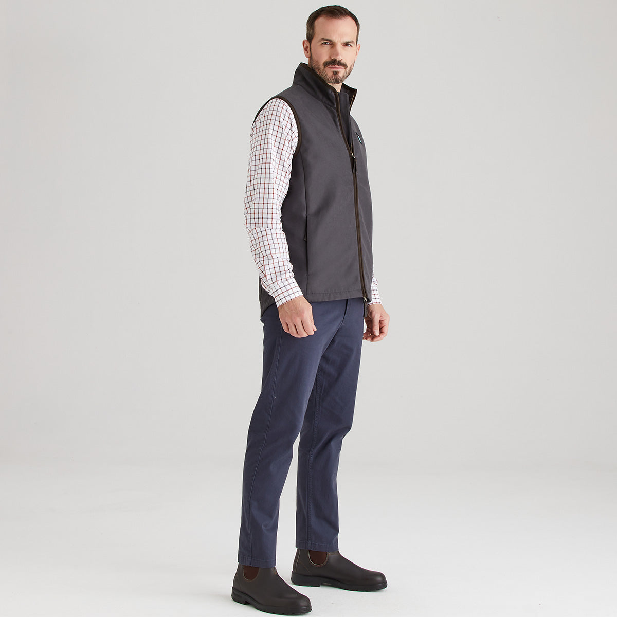 Man wearing a grey vest over a checkered shirt with navy trousers on a light grey background
