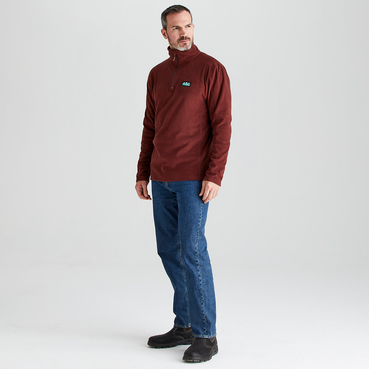 full side image of a man wearing a ridgeline 1/4 zip fleece, blue jeans and a working pair of shoes in a gray backdrop
