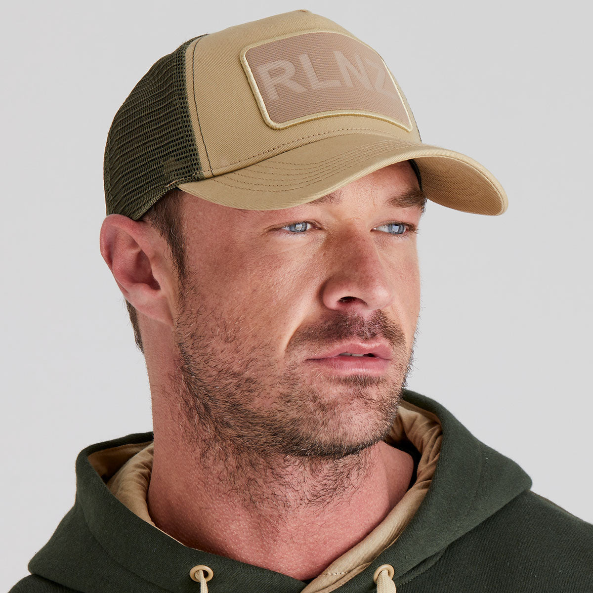 Man wearing a tan cap with a patch and green hoodie on a gray background