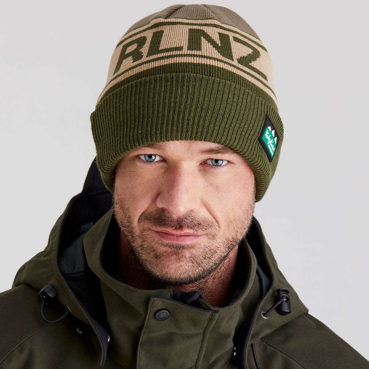 Man wearing a green beanie with 'RLNZ' branding and a matching green jacket on a light gray background.