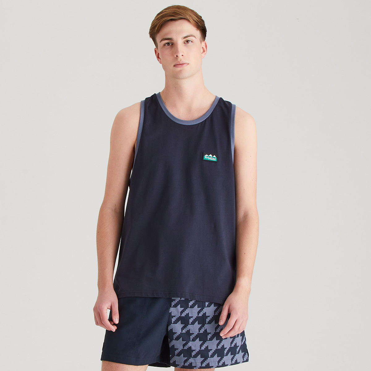 Person wearing a navy tank top with a logo and patterned shorts on a light gray background