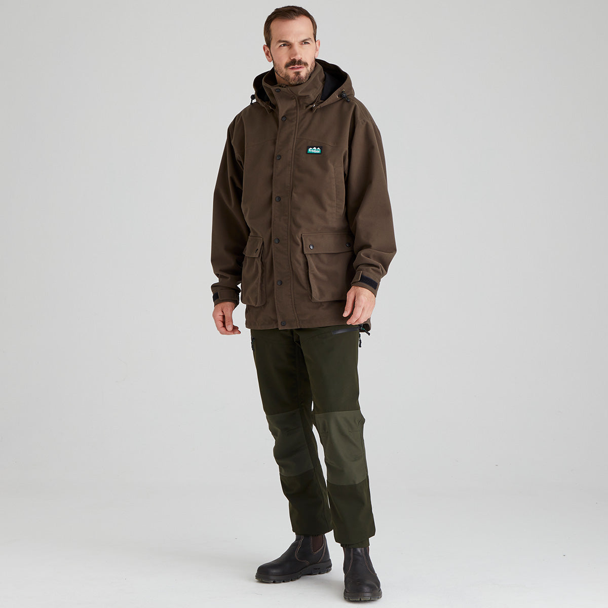 Man wearing a brown waterproof jacket and green pants on a grey background