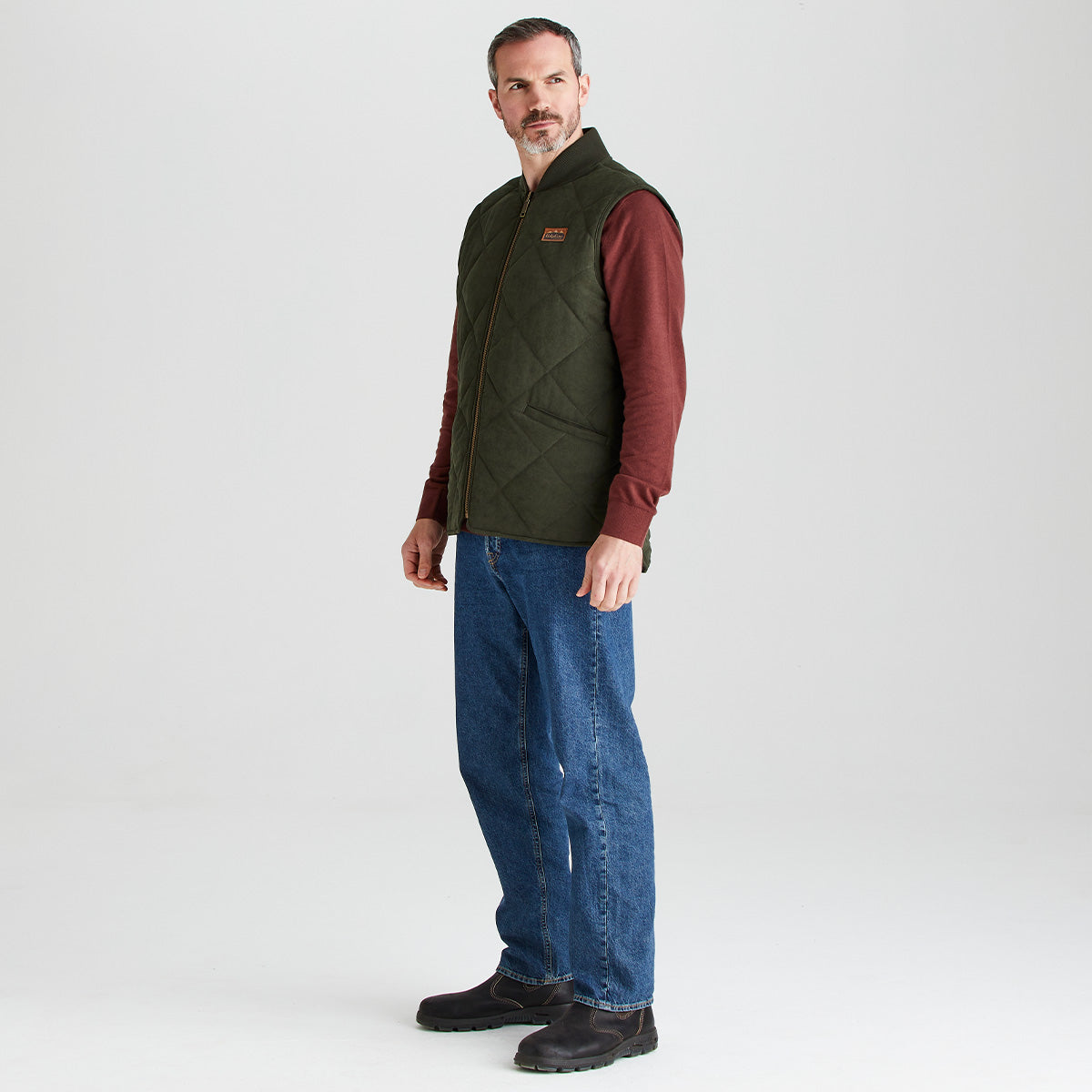 Man wearing a green quilted vest over a red shirt, blue jeans, and black boots on a white background