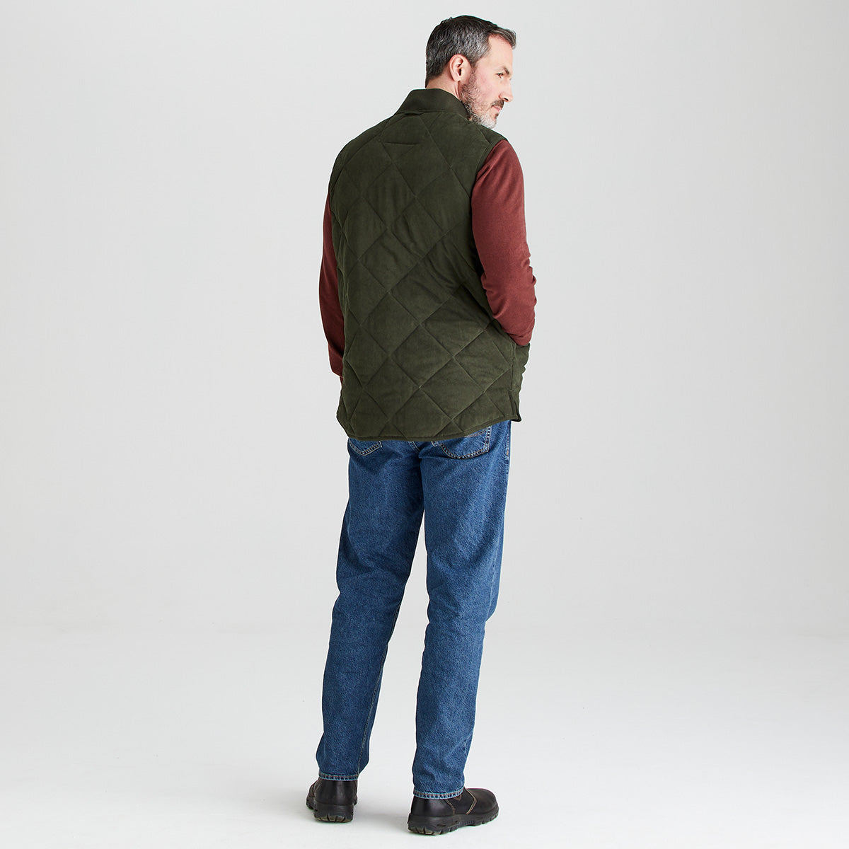 Man wearing a green quilted vest over a red shirt with blue jeans on a white background