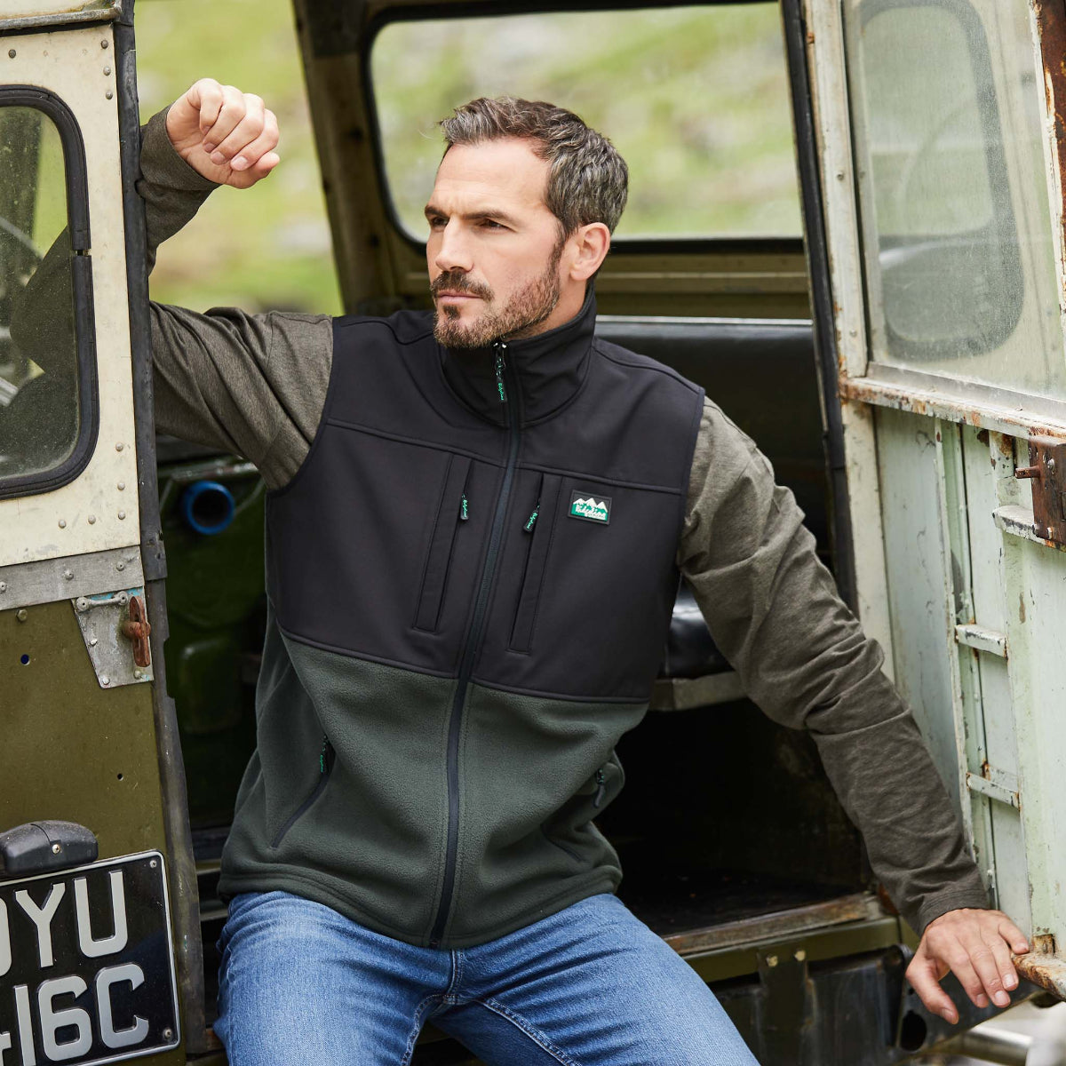 man wearing Two-tone black and green Windproof fleece gilet with a Ridgeline Clothing logo leaning out of the back on a jeep