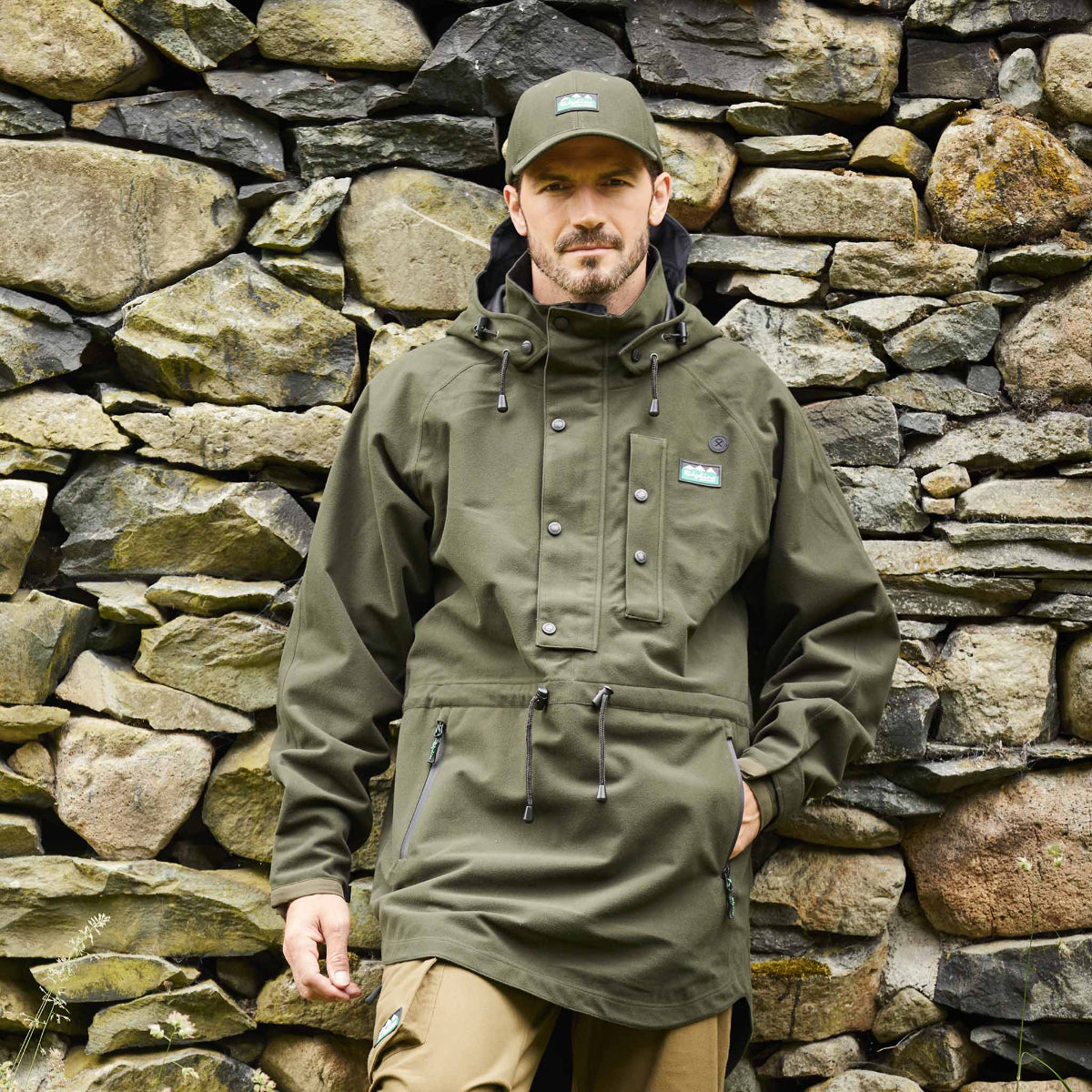 Man wearing a green jacket and cap standing against a stone wall