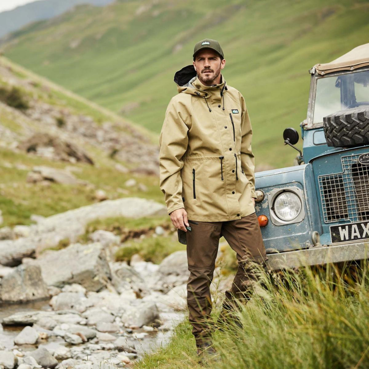 Man in a beige jacket standing next to a vintage Land Rover in a mountainous landscape.