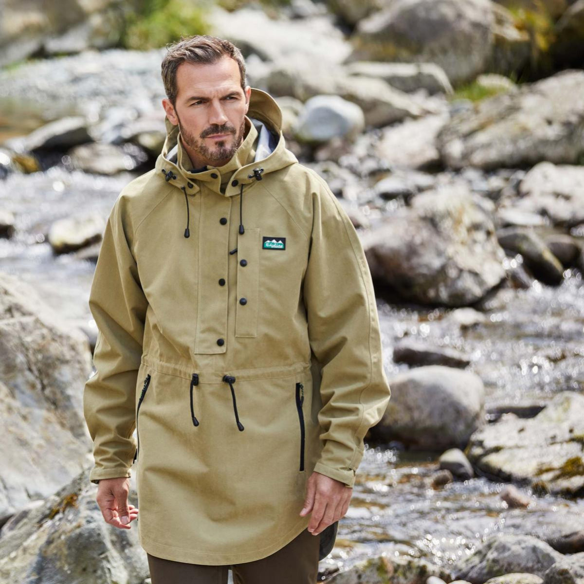 Man wearing a beige raincoat standing by a rocky stream
